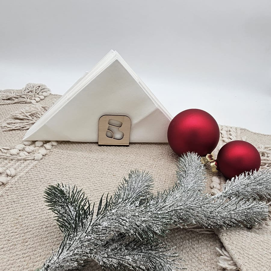 Weihnachtlicher Holz-Serviettenhalter mit Stiefelmotiv, kombiniert mit weißen Servietten, roten Christbaumkugeln und Tannenzweig auf beiger Tischdecke – natürliche Weihnachtsdekoration.