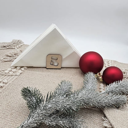 Weihnachtlicher Holz-Serviettenhalter mit Stiefelmotiv, kombiniert mit weißen Servietten, roten Christbaumkugeln und Tannenzweig auf beiger Tischdecke – natürliche Weihnachtsdekoration.