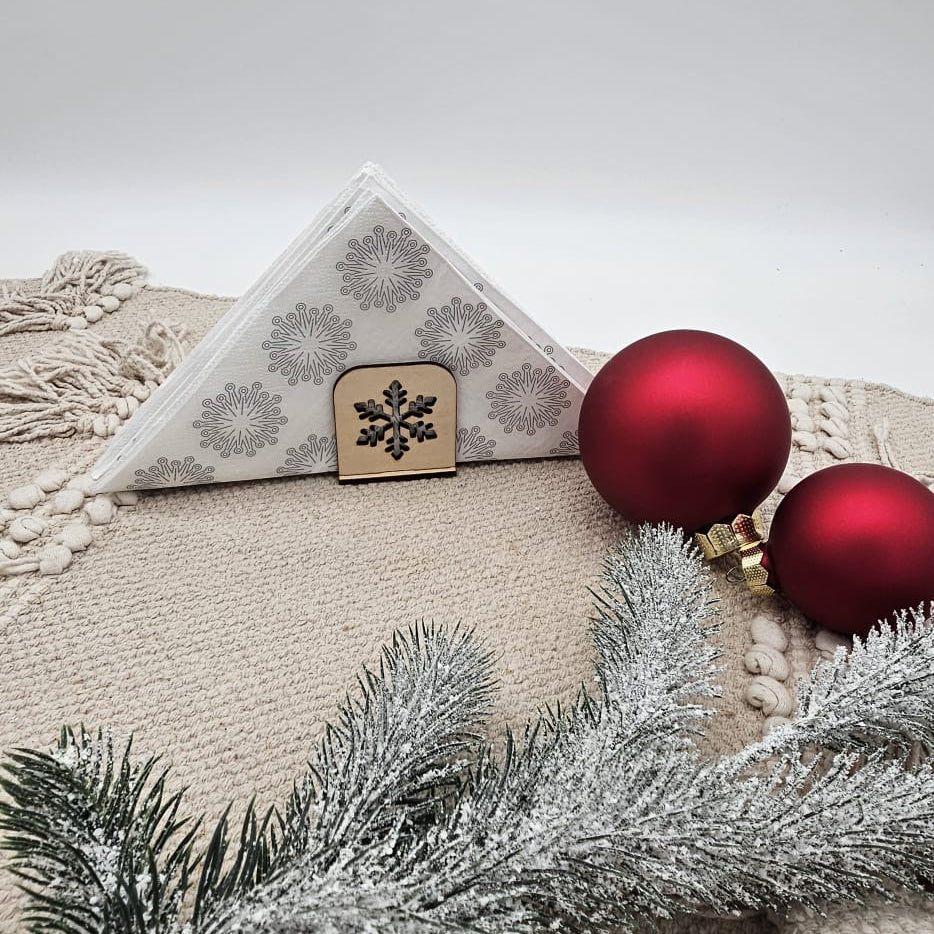 Holz-Serviettenhalter mit Schneeflockenmotiv, kombiniert mit weißen Servietten, roten Weihnachtskugeln und Tannenzweig auf beiger Tischdecke – festliche, natürliche Weihnachtsdekoration.