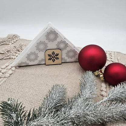 Holz-Serviettenhalter mit Schneeflockenmotiv, kombiniert mit weißen Servietten, roten Weihnachtskugeln und Tannenzweig auf beiger Tischdecke – festliche, natürliche Weihnachtsdekoration.