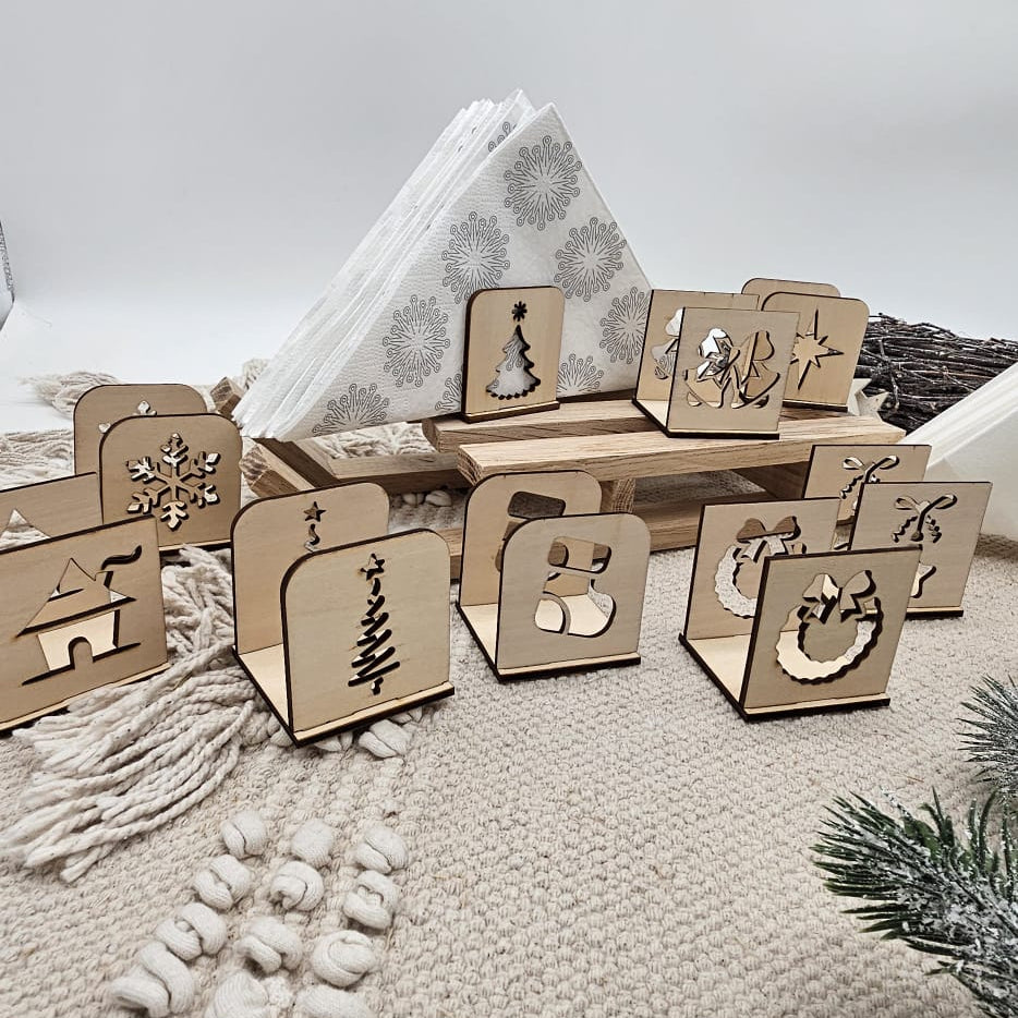 Handgefertigte Holz-Serviettenhalter mit verschiedenen Weihnachtsmotiven – Tannenbaum, Stern, Schneeflocke, Stiefel, Kranz und Haus – auf beiger Tischdecke mit Servietten und Tannenzweig dekorativ angeordnet.