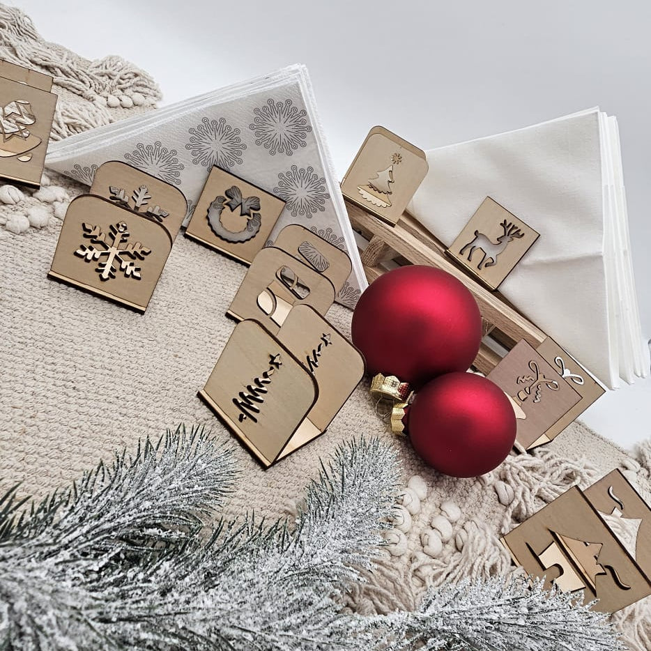 Weihnachtliche Serviettenhalter aus Holz mit verschiedenen Motiven – Tannenbaum, Schneeflocke, Rentier und Kranz – auf beige Tischdecke mit roten Christbaumkugeln und Tannenzweig dekoriert.