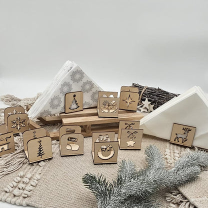 Set aus handgefertigten Holz-Serviettenhaltern mit verschiedenen Weihnachtsmotiven wie Tannenbaum, Stern, Kranz, Rentier und Schneeflocke – arrangiert auf beiger Tischdecke mit Servietten und Tannenzweig im Vordergrund.