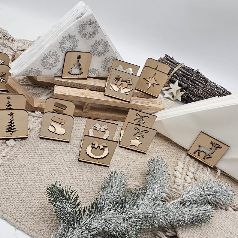 Set aus handgefertigten Holz-Serviettenhaltern mit verschiedenen Weihnachtsmotiven – Tannenbaum, Stern, Geschenk, Kranz und Rentier – auf beiger Tischdecke mit Servietten und Tannenzweig arrangiert.